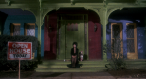 A Gothic-styled woman seated on the steps leading to a Gothic-styled house.