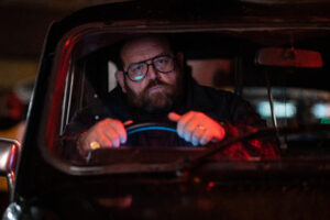 Image looking through window of a black cab at a bearded man wearing glasses