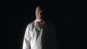 Nicholas Vince wearing white clothing against a black background